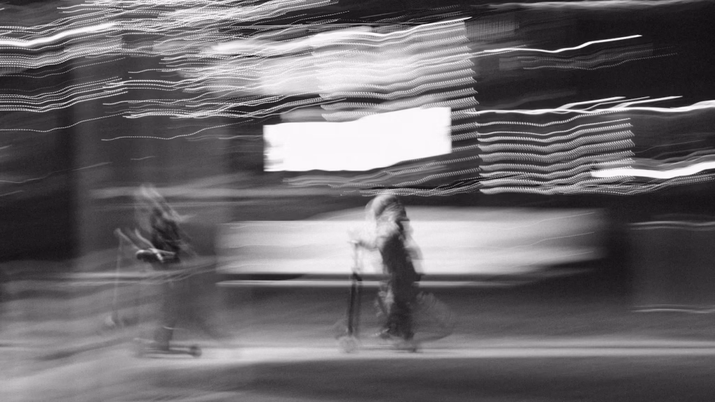 Abstract black and white long-exposure photograph showing blurred pedestrians and light trails at night, evoking uncertainty and perception.
